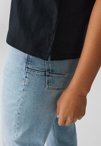 T-shirt nera in cotone con spacco laterale, abbinata a jeans denim azzurri chiari con tasca posteriore e texture strappata, tenuta vicino all'anca.