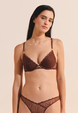 Jeune femme aux longs cheveux foncés portant un soutien-gorge et une culotte en dentelle marron assortis, debout devant un fond beige uni.