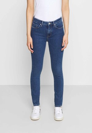 Slim-fit blue jeans worn by person in white top and white sneakers, standing against plain light background.