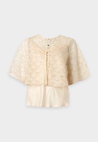 Veste boléro en dentelle beige à sequins avec larges manches courtes, portée sur un top beige uni sans manches.