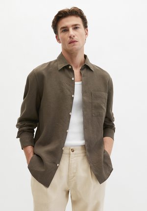 Junger Mann mit Händen in den Taschen, der ein aufgeknöpftes olivgrünes Hemd über einem weißen Tanktop und beigen Hosen vor neutralem Hintergrund trägt.