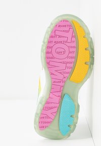 Semelle de basket avec bords translucides, partie rose avec le texte « TOMMY JEANS », zones d’accroche jaunes et bleues, et grandes lettres embossées « TOMMY ».