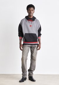Sweat à capuche gris et noir avec des accents rouges, poche frontale et patch logo. Le modèle porte un jean gris délavé et des bottes noires.