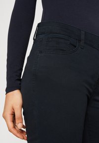 Gros plan d'une personne portant un pantalon sombre taille haute avec une petite poche avant, visible de la taille jusqu'au milieu de la cuisse, la main reposant à proximité.