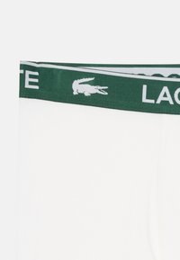Boxer bianchi con un elastico verde in vita, con la scritta "LACOSTE" e un logo coccodrillo bianco. Texture liscia.