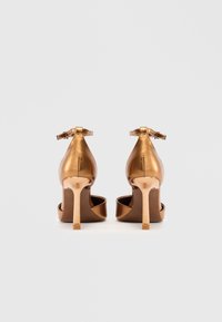 Brončane visoke potpetice s sjajnom površinom, trakom oko gležnja i suženim potpeticama. Dizajn s otvorenim prstima s glatkom teksturom.