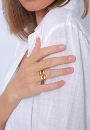 Guldbelagt metallring med en organisk, abstrakt form. Båret på en hånd, der hviler på et hvidt, tekstureret tøj. Glat overfladefinish.