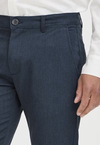 Marina skräddarsydda byxor med knappstängning, enhetlig textur och sneda sidofickor, ihop med en vit skjorta med krage.