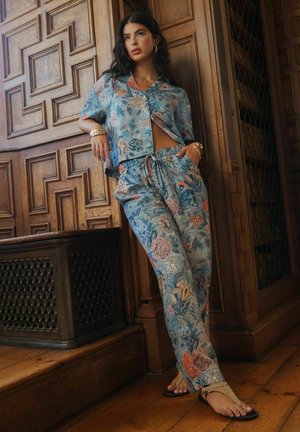 Frau lehnt an einer holzvertäfelten Wand, trägt ein passendes blaues Hemd und Hose mit floralem Muster, Sandalen, Goldschmuck und hat die Haare offen.