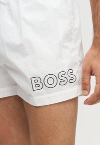 Short blanc avec texte "BOSS" en contour noir sur la jambe gauche, porté par une personne avec une main visible et le haut de la cuisse.
