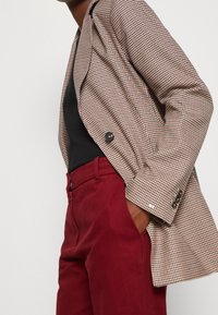 Persona con un blazer estampado sobre una camiseta negra, con una mano en el bolsillo de unos pantalones de tiro alto de color rojo oscuro, frente a un fondo sencillo.