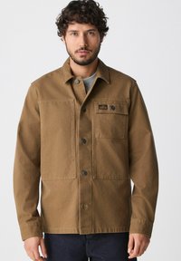 Brown cotton jacket with two front pockets, button closure, and a small label. Features a collar and buttoned cuffs, textured fabric.