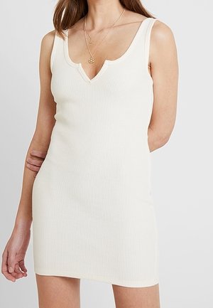 Mouwloze, geribbelde witte minijurk met een V-hals, gedragen door een persoon met gelaagde gouden kettingen.