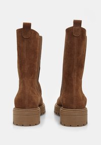 Botine maro din suede, cu glezne drepte, suprafețe texturate și tălpi groase din cauciuc bej. Bucles în partea de sus pentru o îmbrăcare ușoară.