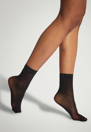 Gambe in calze nere trasparenti alla caviglia con fasce decorative di strass, un piede leggermente sollevato su uno sfondo chiaro e semplice.