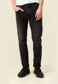 Hombre con jeans negros desgastados, una mano en el bolsillo y zapatos negros de cordones, sobre un fondo beige.