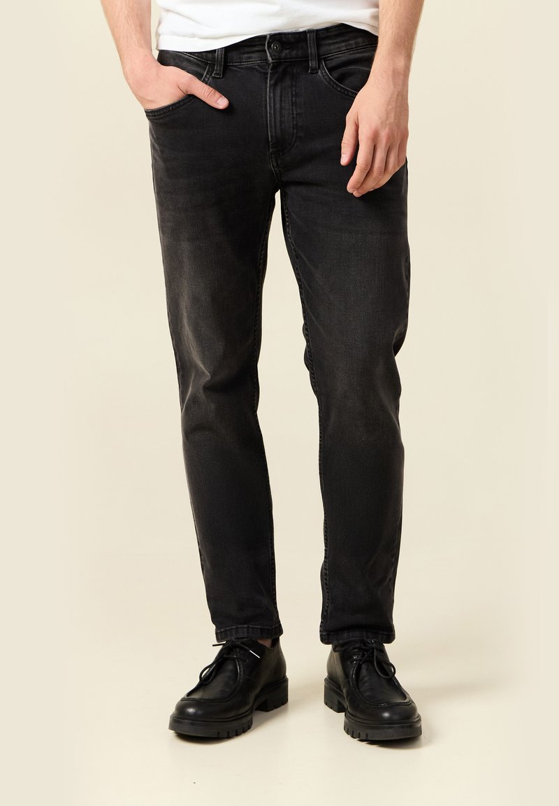 Hombre con jeans negros desgastados, una mano en el bolsillo y zapatos negros de cordones, sobre un fondo beige.