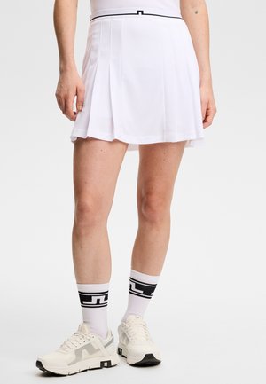 Personne portant une jupe de tennis plissée blanche, des chaussettes montantes blanches à motifs noirs, et des baskets de sport blanches, debout sur un fond uni.
