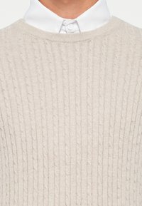 Lys beige strikket genser med kabelmønster, rund hals og ribbestrikket nederkant, båret over en hvit skjorte med krage.
