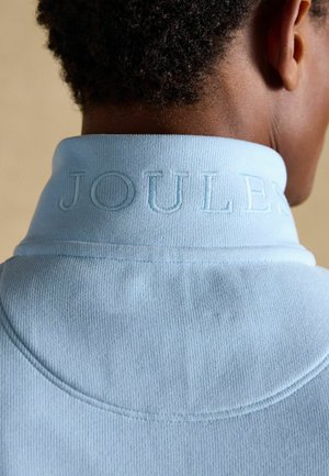 Tagantvaade inimesest, kes kannab helesinist kõrge kaelusega rõivast, millel kaelusel on reljeefne tekst "JOULES", näidates lühikest lokkis juustest ja kaela.