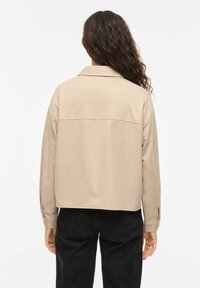 Bēša, pārizmēra krekls ar apkakli, garām piedurknēm un ar pogām aizpogājamiem aprocēm. Gluda tekstūra, cropped dizains un detaļa aizmugures yoke daļā.