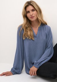 Blå, langærmet bluse med V-hals, tekstureret stof og samlede manchetter. Båret over sorte bukser, mod en neutral baggrund.