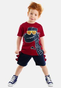 Rotes Baumwoll-T-Shirt mit einem Dinosaurier-Motiv und dem Text "Cool Boy". Kombiniert mit blauen Shorts mit einem Rüschensaum und weißen Sneakers.