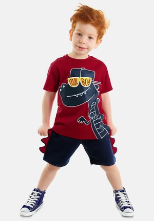 Rotes Baumwoll-T-Shirt mit einem Dinosaurier-Motiv und dem Text "Cool Boy". Kombiniert mit blauen Shorts mit einem Rüschensaum und weißen Sneakers.