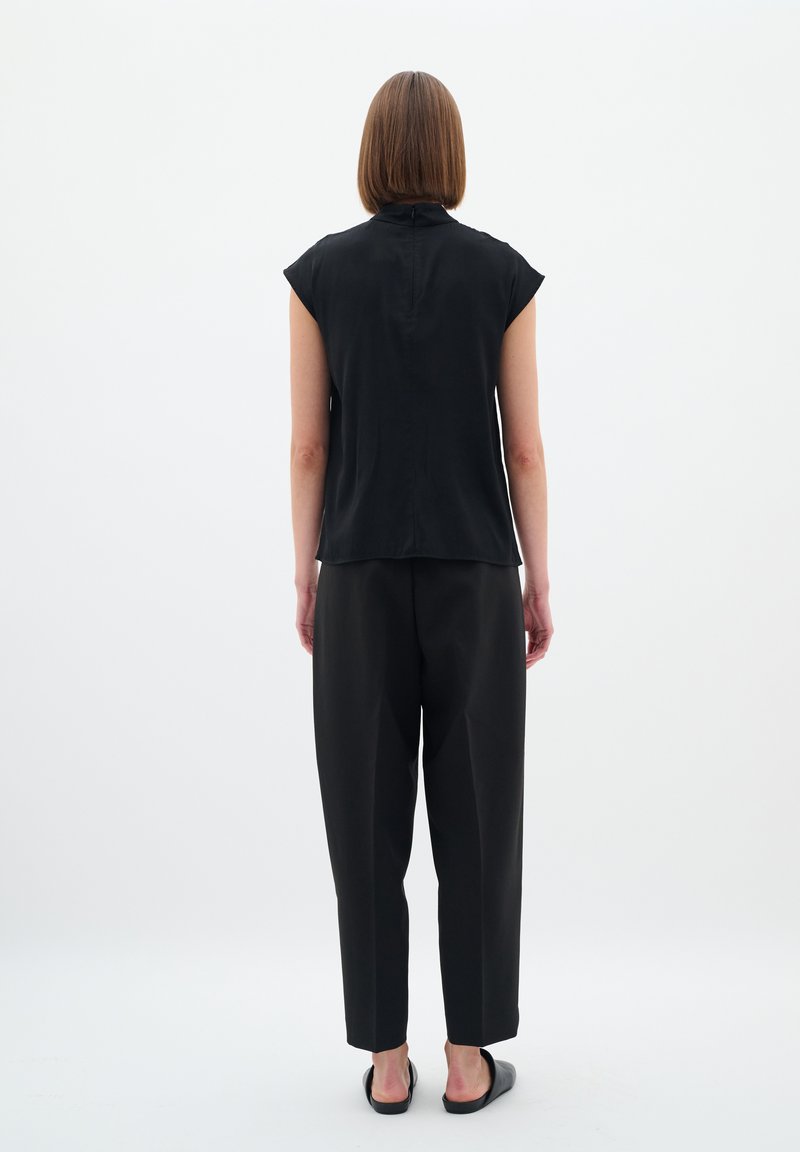 Schwarzes, kurzärmeliges Oberteil mit hohem Kragen und glatter Textur, kombiniert mit lockeren schwarzen Hosen, von hinten vor einem weißen Hintergrund betrachtet.