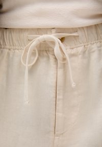 Beige housut, joissa on joustava vyötärö ja kangassolki. Pehmeä rakenne, sivutaskut ja materiaalissa on hieno kiilto.