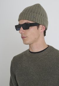 Uomo che indossa un berretto lavorato a maglia verde oliva, un maglione a coste abbinate e occhiali da sole neri rettangolari, rivolto a sinistra su uno sfondo semplice.