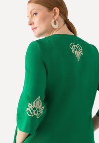 Grüne Bluse mit 3/4-Ärmeln, featuring beige dekorativer Stickerei an Ärmeln und Rücken. Glatter, leichter Stoff mit lässiger Passform.