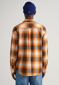 Camicia di flanella a scacchi arancione, marrone e grigia, con maniche lunghe, polsini a bottone e orlo curvo. Berretto di maglia blu.