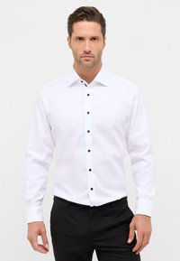 Man met gestyled haar, gekleed in een wit overhemd met lange mouwen en zwarte knopen, en zwarte broek, staande tegen een eenvoudige lichte achtergrond.