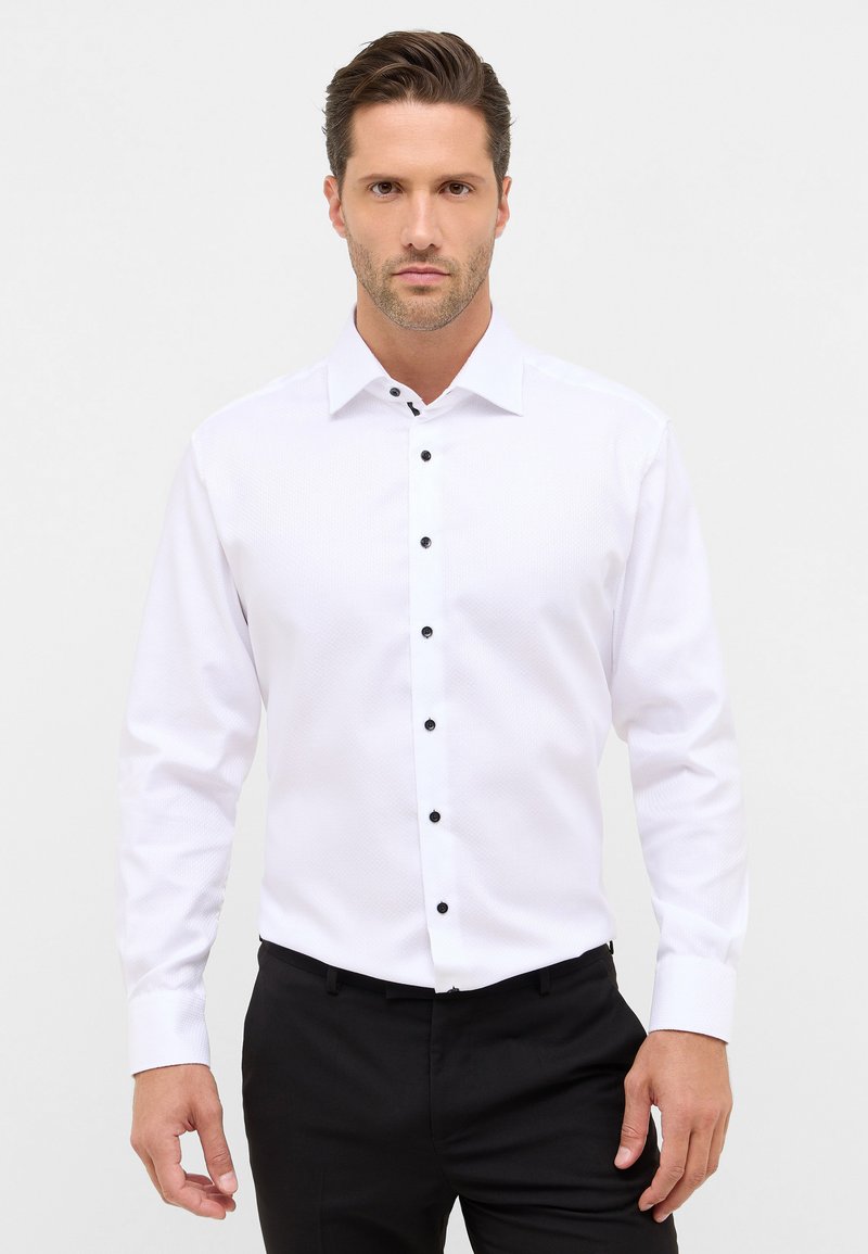 Man met gestyled haar, gekleed in een wit overhemd met lange mouwen en zwarte knopen, en zwarte broek, staande tegen een eenvoudige lichte achtergrond.