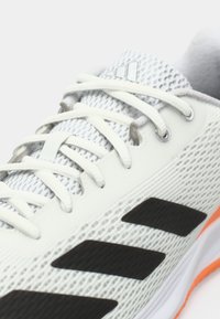 Chaussure de sport blanche et grise avec des lacets blancs, des rayures noires sur le côté et une semelle bordée d'orange.