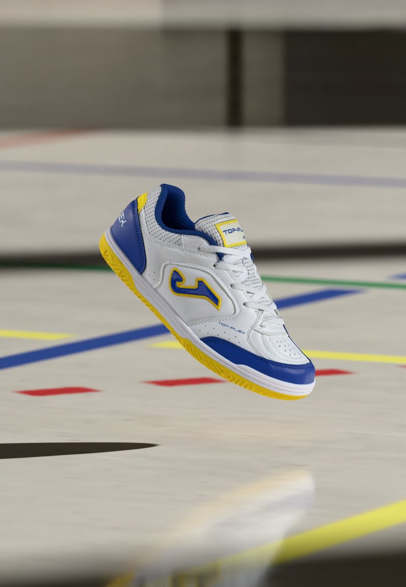 Chaussure de sport d’intérieur blanche, bleue et jaune suspendue au-dessus d’un sol de gymnase avec des lignes et des marquages colorés.