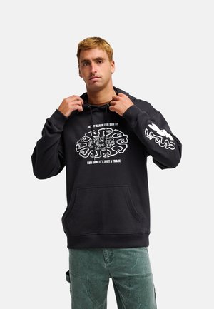Mann mit kurzen blonden Haaren trägt einen schwarzen Hoodie mit weißem Grafikdesign und Text, richtet die Kapuzenbänder und steht vor einem schlichten Hintergrund.