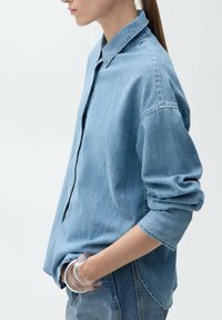Hellblaues Jeanshemd mit Knopfleiste, lockerer Passform und langen Ärmeln, die an den Manschetten hochgerollt sind. Glatte Textur und klassisches Kragendetail.