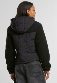 Schwarze Kapuzenjacke aus Sherpa- und Steppstoff. Verfügt über strukturierte Ärmel und Rücken, lockere Passform und ein angenehmes Tragegefühl.