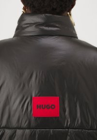 Svart vadderad jacka med hög krage och en framträdande röd rektangulär lapp märkt "HUGO" på baksidan. Slät, glänsande yta.