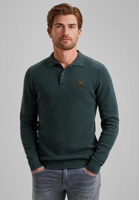 Suéter polo de manga larga en verde oscuro con un tejido texturizado, tapeta de tres botones y un pequeño parche de logo cuadrado en el pecho.