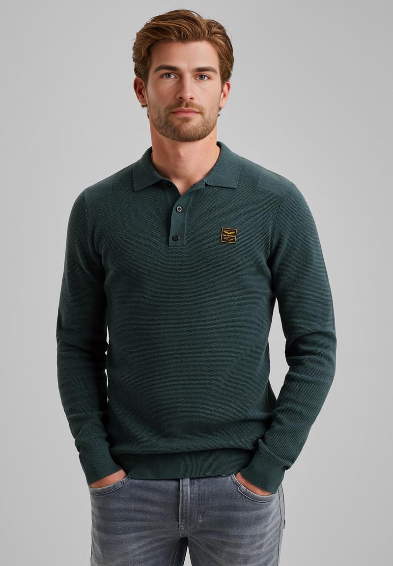 Suéter polo de manga larga en verde oscuro con un tejido texturizado, tapeta de tres botones y un pequeño parche de logo cuadrado en el pecho.