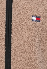 La giacca in pile beige peloso presenta una cerniera nera e una piccola toppa con il logo "TOMMY JEANS" in blu, rosso e bianco sul davanti.
