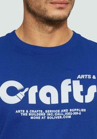 Blauwe t-shirt met grote witte tekst 'Arts & Crafts' en een verfkwast grafiek. Gemaakt van katoen met een ronde hals.