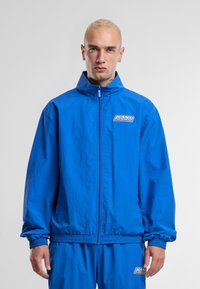 Blue lightweight windbreaker with a zip front, stand collar, elastic cuffs, and waistband; features a logo patch on the left chest.