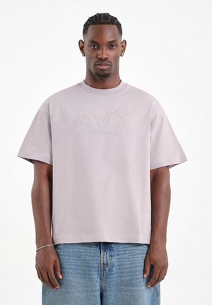 Hombre con cabello trenzado, vistiendo una camiseta oversized de color lila claro con el logo "AVIREX DEPT." y jeans azules, de pie frente a un fondo blanco.