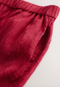 Shorts en tissu rouge froissé avec une ceinture élastique. La texture est douce et légère, avec un design décontracté et fluide.