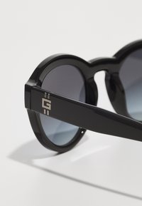 Ochelari de soare negri supradimensionați cu lentile gri degradate, formă rotundă, ramă din plastic lucios, având un logo metalic pe lateral.