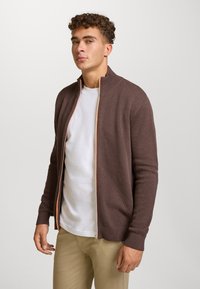 Brun teksturert cardigan med glidelås og høy krage, med kontrasterende oransje glidelås. Brukes over en hvit skjorte og kombinert med lyse bukser.